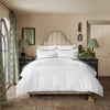 A neatly made bed features the John Robshaw Anketi Natural Border Organic Duvet, crafted from 300 thread count organic cotton, and is adorned with patterned pillows. It is centered between two bedside tables with lamps in a cozy, well-lit bedroom featuring rustic decor and brick flooring.