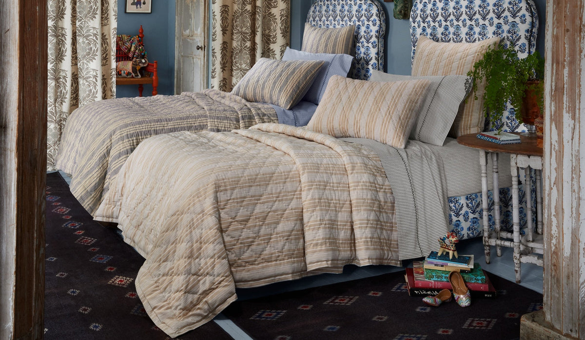 Two twin beds with patterned bedding and pillows are side by side in a cozy bedroom. A small table with a plant and books, slippers, and additional items are on the dark patterned rug beside the beds.