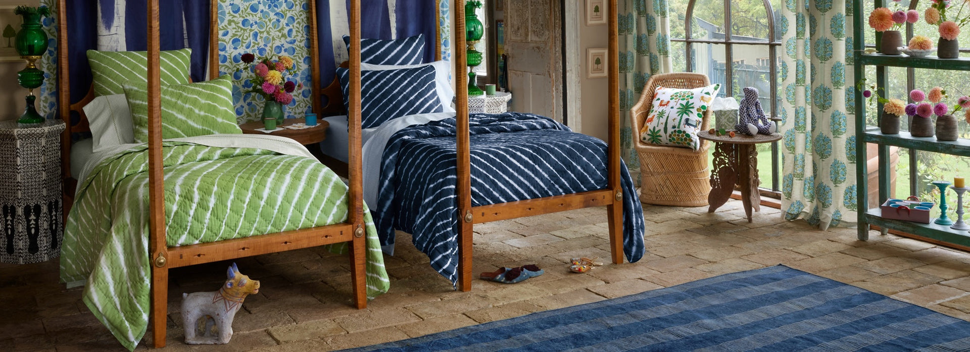 Two wooden canopy beds with green and blue striped bedding in a bright, eclectic bedroom with plants, colorful decor, a wicker chair, and large windows letting in natural light.