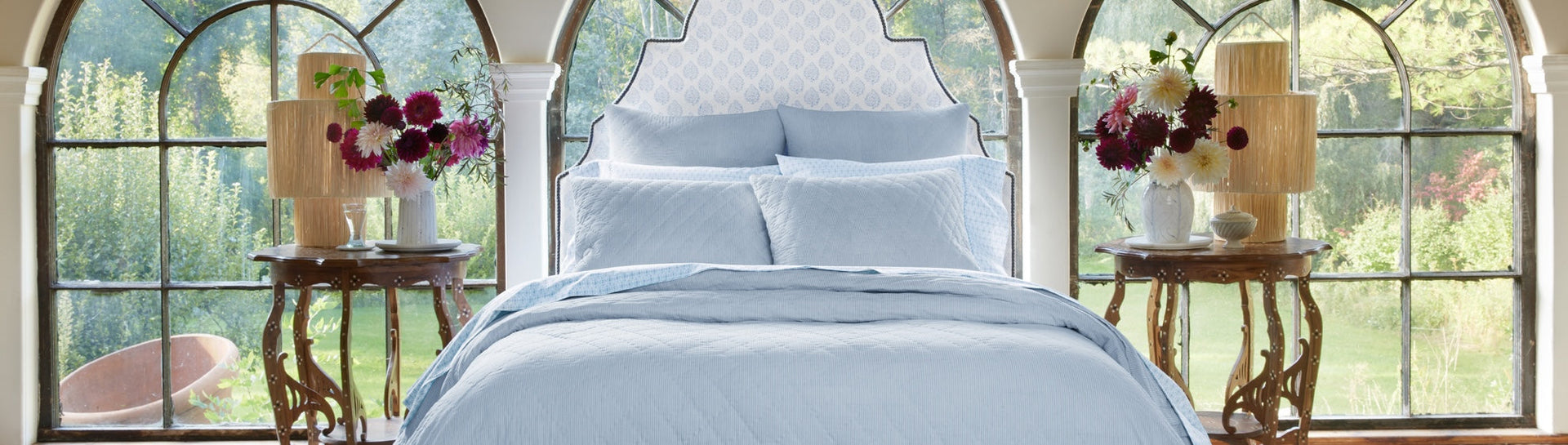 A neatly made bed with light blue bedding sits between two wooden side tables topped with lamps and flower vases, with large arched windows showing a green garden view in the background.