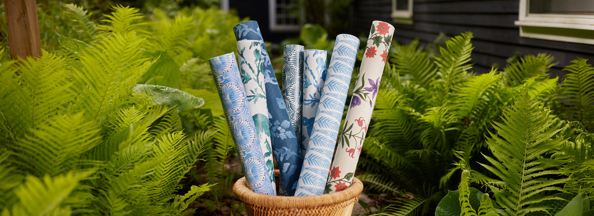 A basket filled with various rolls of patterned wallpaper, featuring blue, white, and floral prints, is placed outdoors among green ferns.