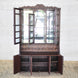 The Nisbat Teak Cabinet by John Robshaw, featuring glass-paneled mirrored doors above and three open storage compartments below with elegant hand-carved detailing, stands against a white brick wall on a woven rug.