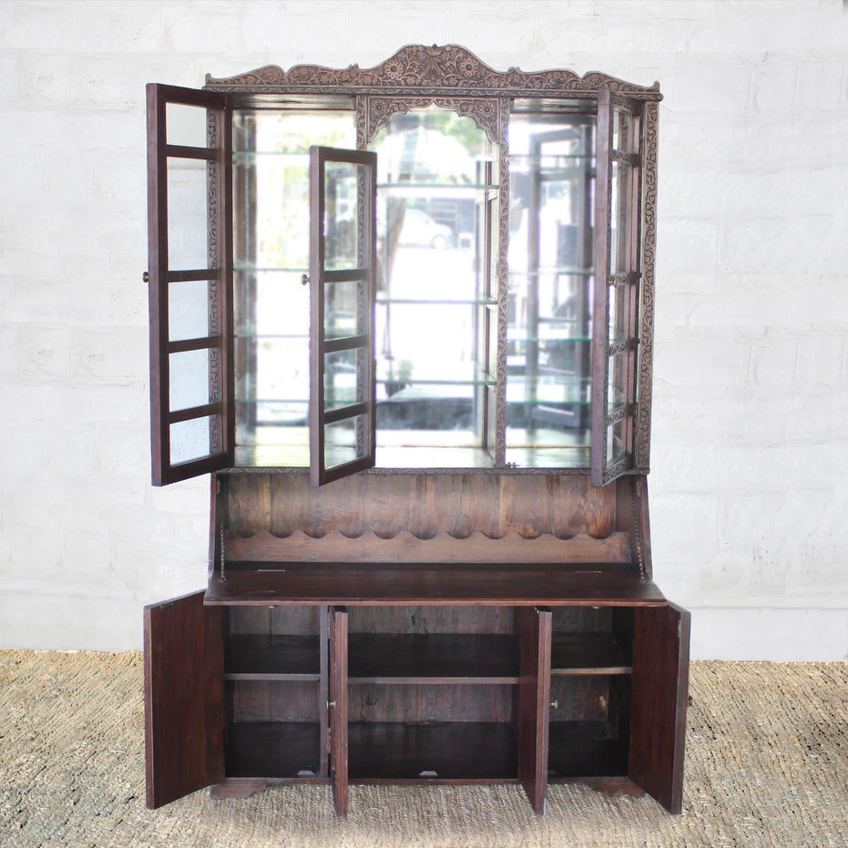 The Nisbat Teak Cabinet by John Robshaw, featuring glass-paneled mirrored doors above and three open storage compartments below with elegant hand-carved detailing, stands against a white brick wall on a woven rug.