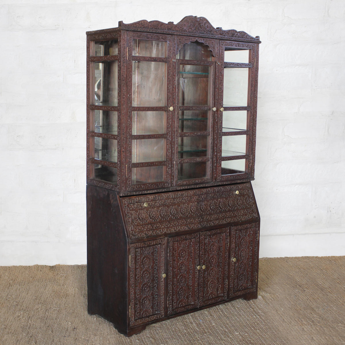 The Nisbat Teak Cabinet by John Robshaw features glass-paneled doors on the upper section, exquisite hand-carved detailing, and lower storage compartments, shown here against a white brick wall.