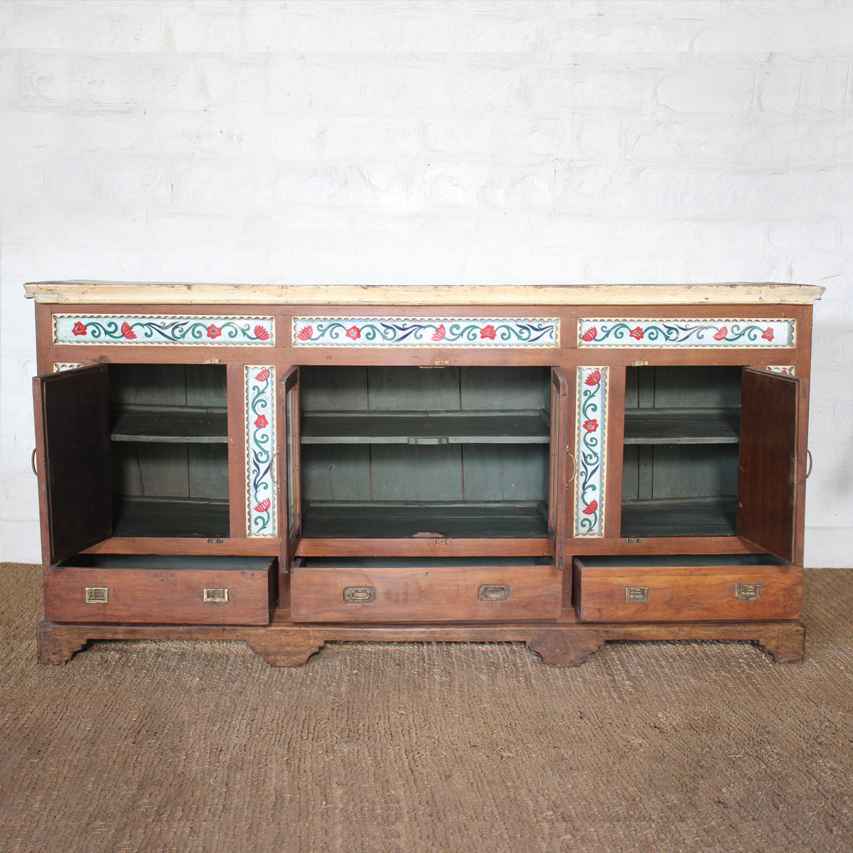 The Gulab Side Board by John Robshaw is a vintage teak sideboard featuring three open cabinet doors and three drawers below, adorned with a painted floral design along the upper front and accented by elegant floral tile insets for added charm.