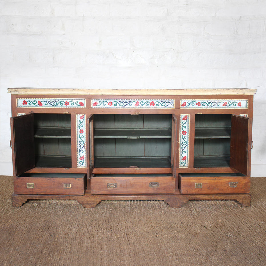 The Gulab Side Board by John Robshaw is a vintage teak sideboard featuring three open cabinet doors and three drawers below, adorned with a painted floral design along the upper front and accented by elegant floral tile insets for added charm.