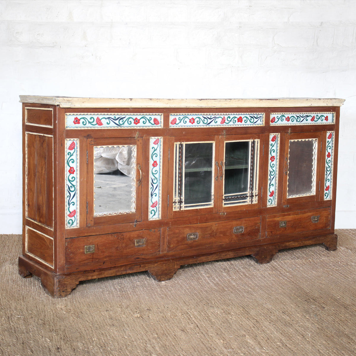 The Gulab Side Board by John Robshaw, featuring four drawers, two mirrored cabinet doors, and floral painted decorative trim, is set against a white brick wall on a woven floor rug.