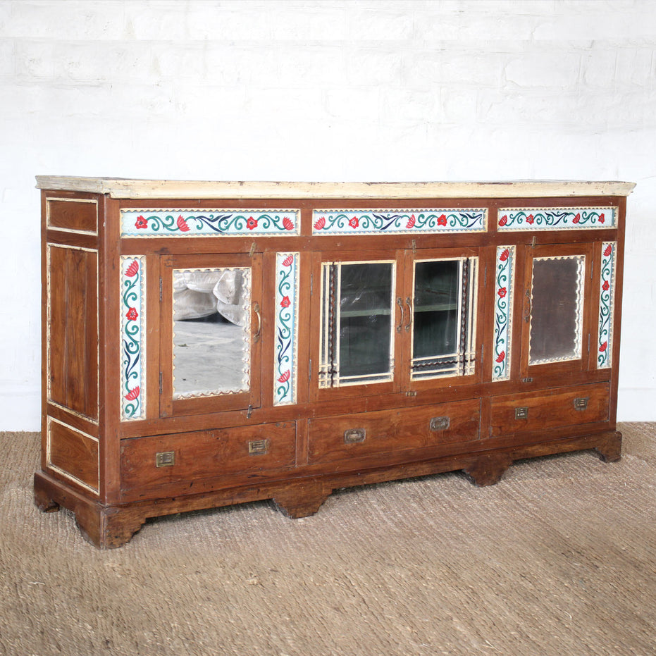 The Gulab Side Board by John Robshaw, featuring four drawers, two mirrored cabinet doors, and floral painted decorative trim, is set against a white brick wall on a woven floor rug.