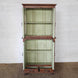The Sunehri Teak Cabinet by John Robshaw features open green-painted and arched glass doors, two shelves, and two intricately carved drawers with sunburst panels at the bottom. It stands on a woven rug against a white brick wall.