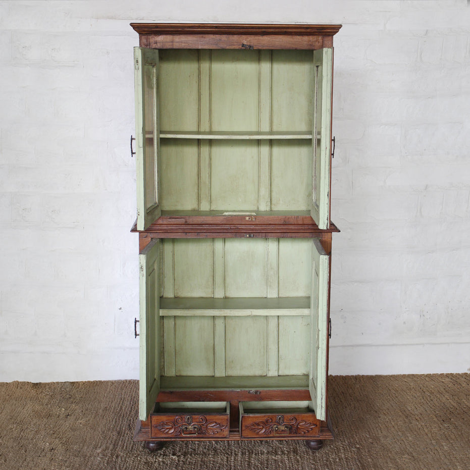 The Sunehri Teak Cabinet by John Robshaw features open green-painted and arched glass doors, two shelves, and two intricately carved drawers with sunburst panels at the bottom. It stands on a woven rug against a white brick wall.