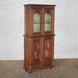 The Sunehri Teak Cabinet by John Robshaw showcases arched glass doors on the upper section and intricately carved sunburst panels on the lower doors, standing tall on a textured brown floor against a white brick wall.