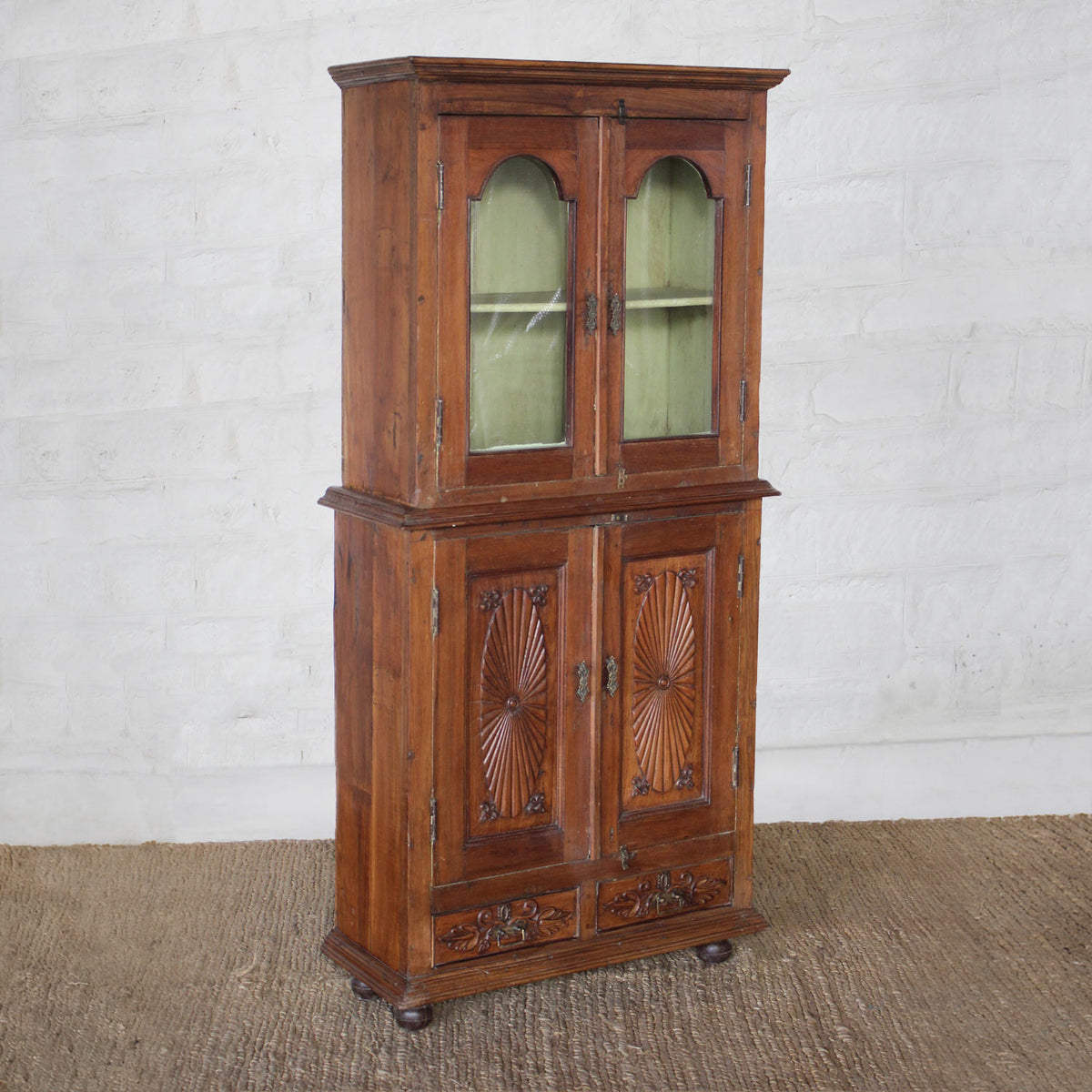 The Sunehri Teak Cabinet by John Robshaw showcases arched glass doors on the upper section and intricately carved sunburst panels on the lower doors, standing tall on a textured brown floor against a white brick wall.