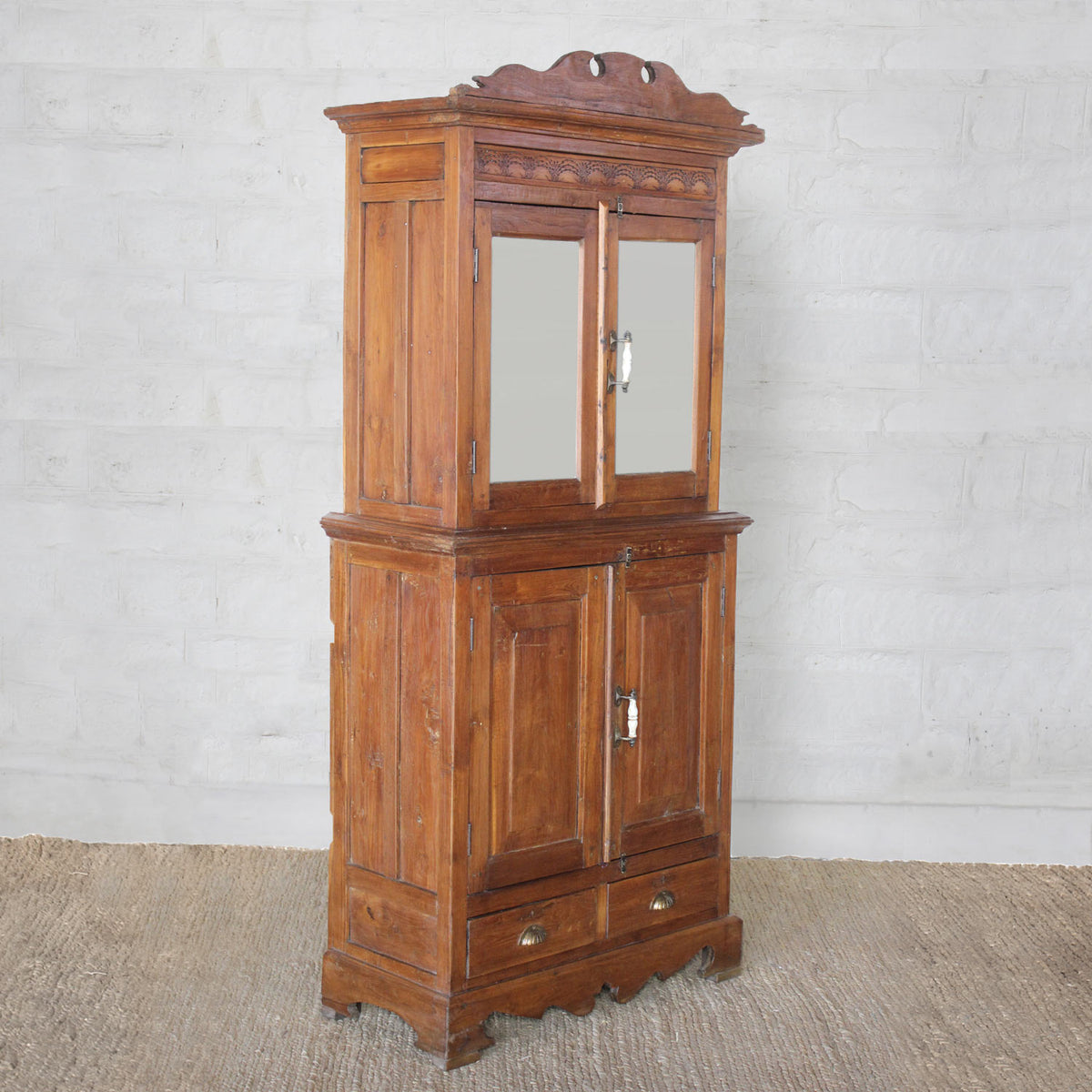 The Sajawat Teak Cabinet by John Robshaw is a tall vintage-inspired piece featuring an ornate top, mirrored upper doors, solid lower doors, and two bottom drawers. Set on a woven rug against a light-colored brick wall, this elegant storage cabinet perfectly blends style and function, making it an exceptional choice for classic interiors.