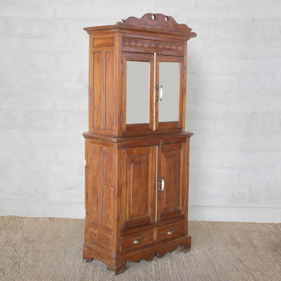 The Sajawat Teak Cabinet by John Robshaw is a tall vintage-inspired piece featuring an ornate top, mirrored upper doors, solid lower doors, and two bottom drawers. Set on a woven rug against a light-colored brick wall, this elegant storage cabinet perfectly blends style and function, making it an exceptional choice for classic interiors.
