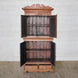 The Sajawat Teak Cabinet by John Robshaw stands on a woven rug against a white brick wall, featuring an ornate top and open double doors that reveal spacious storage cabinets and shelves inside. Two drawers with brass handles at the bottom add both charm and practicality.