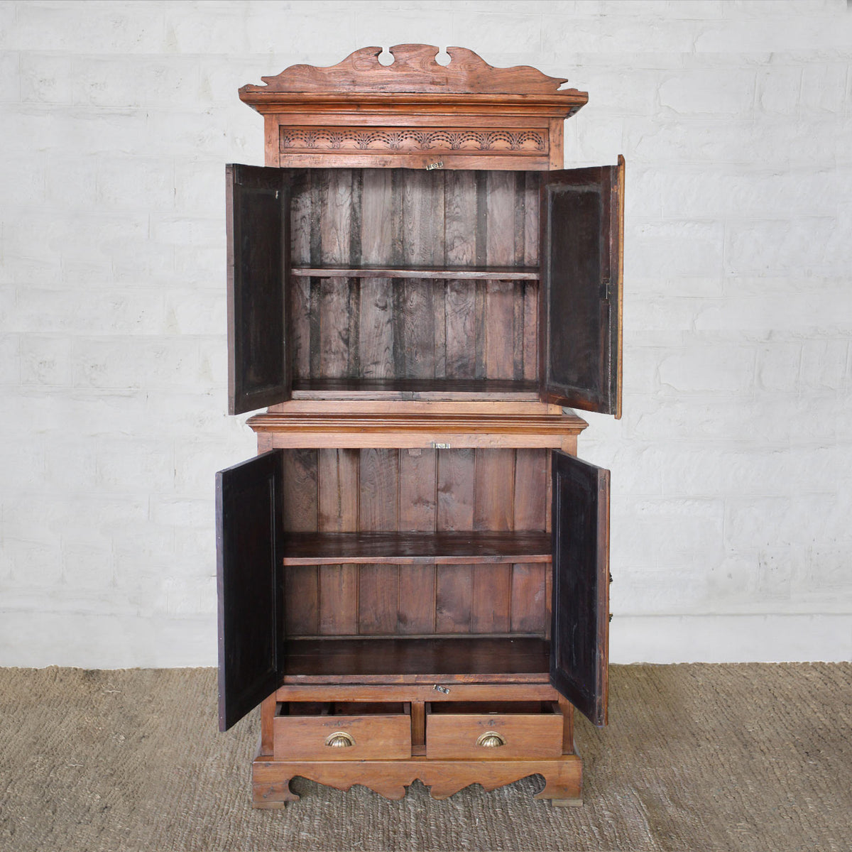 The Sajawat Teak Cabinet by John Robshaw stands on a woven rug against a white brick wall, featuring an ornate top and open double doors that reveal spacious storage cabinets and shelves inside. Two drawers with brass handles at the bottom add both charm and practicality.