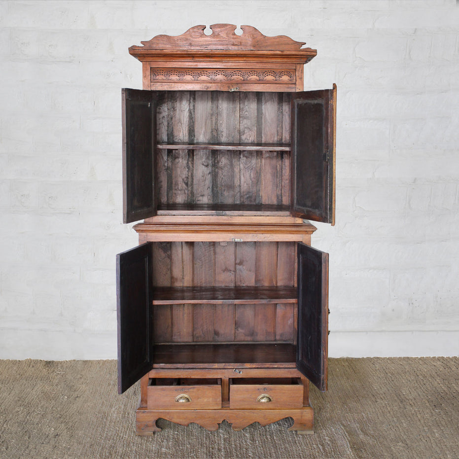 The Sajawat Teak Cabinet by John Robshaw stands on a woven rug against a white brick wall, featuring an ornate top and open double doors that reveal spacious storage cabinets and shelves inside. Two drawers with brass handles at the bottom add both charm and practicality.