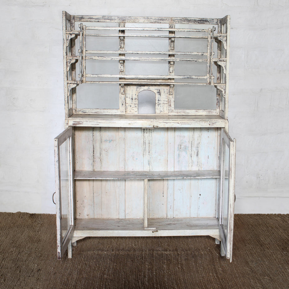 The Saahil Wooden Sideboard by John Robshaw, finished in a charming distressed whitewash, features open upper shelving, elegant glass-front storage panels, and lower cabinet doors that reveal spacious interior shelves—beautifully set against a crisp white wall.