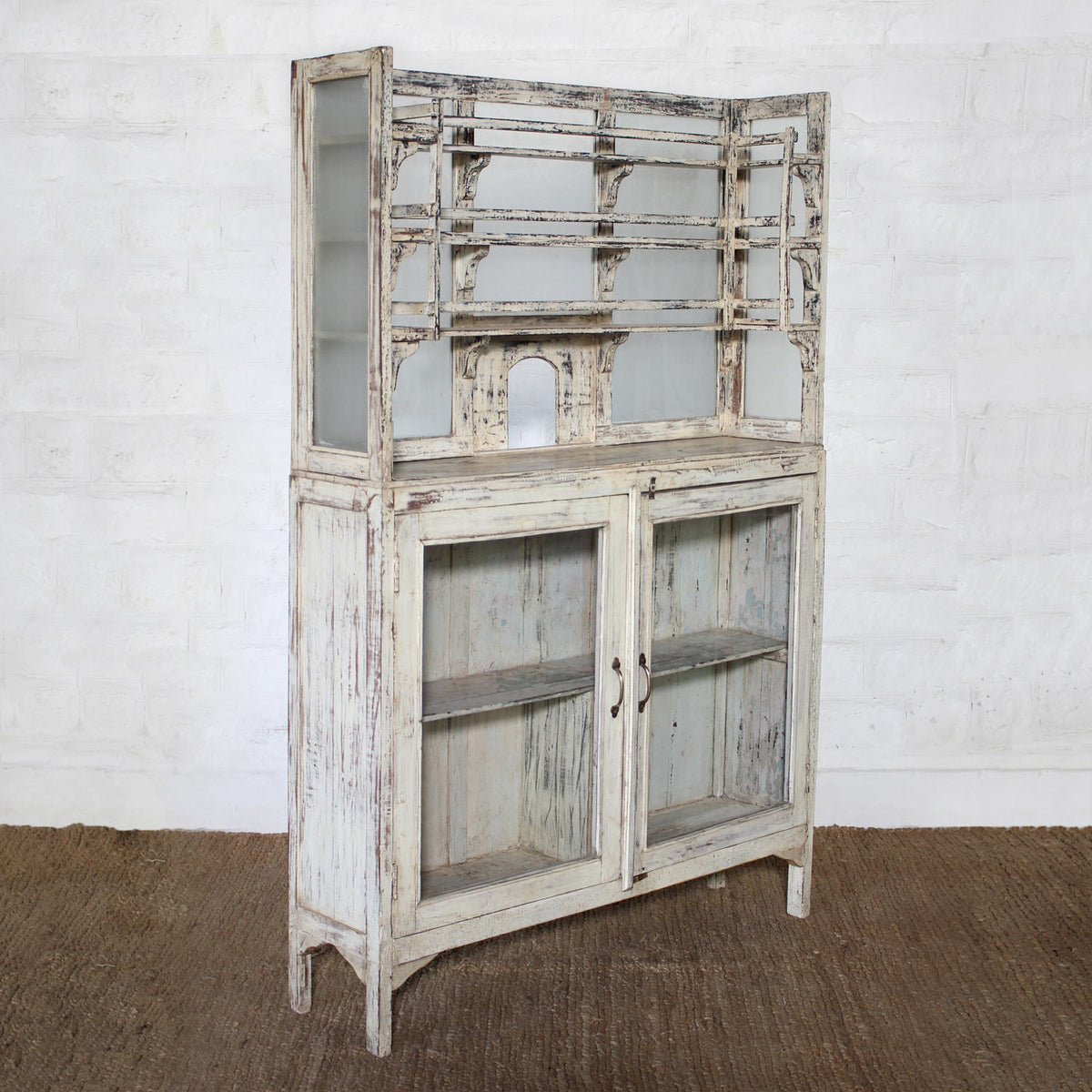 The Saahil Wooden Sideboard by John Robshaw features a distressed whitewashed finish, rustic weathered look, open shelving on top, and glass doors on the lower section for elegant glass-front storage. Shown here set against a white brick wall on a brown rug.