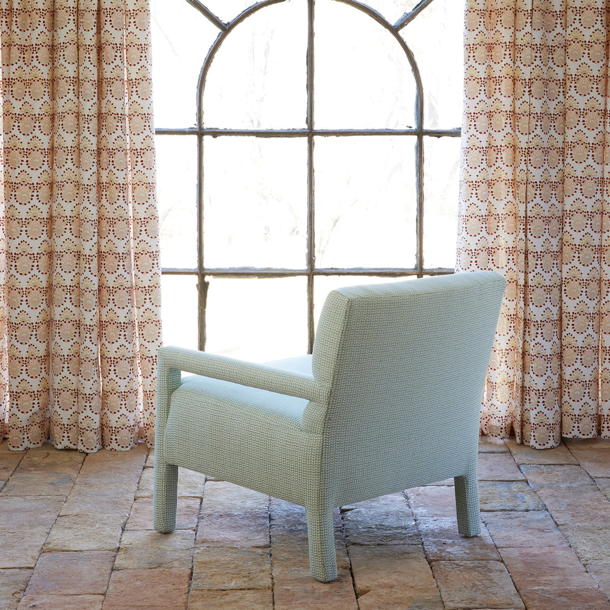 A John Robshaw Square Chair in Aleppo Sage Performance Fabric is upholstered in a light blue tone and positioned on a tiled floor, facing a large arched window with patterned beige curtains.
