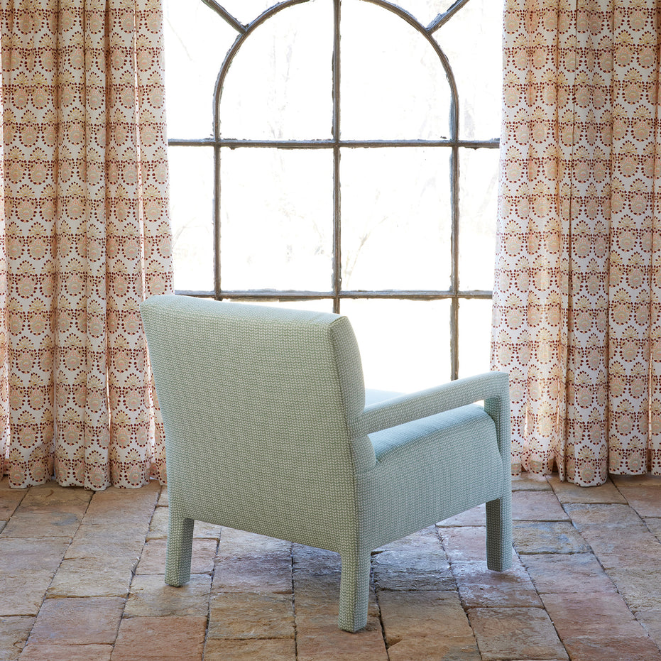 A Square Chair in Aleppo Sage by John Robshaw, featuring custom upholstery, sits on a stone floor in front of a large arched window with patterned peach and white curtains, with natural light streaming in.