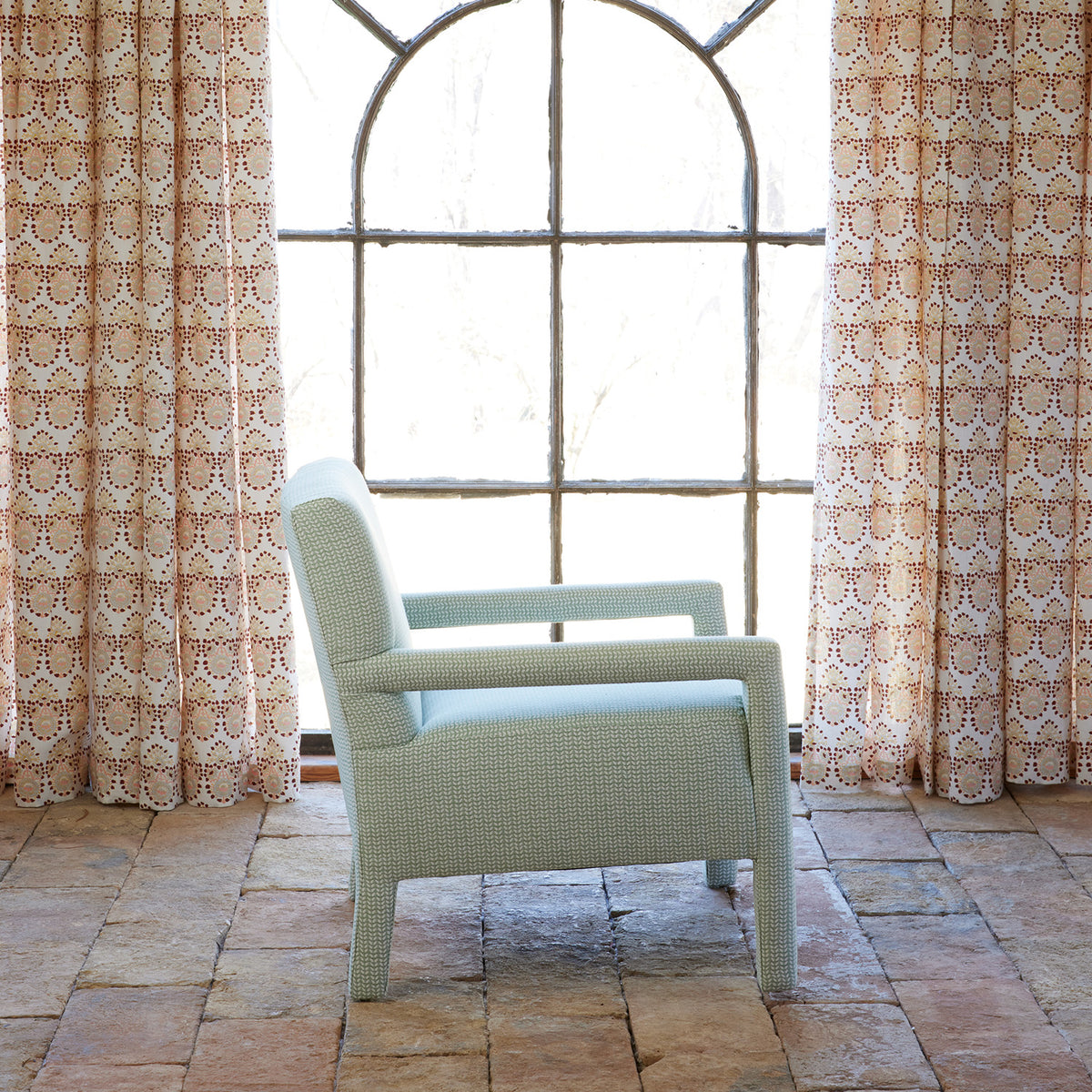 A Square Chair in Aleppo Sage by John Robshaw sits on a tiled floor in front of a large arched window with patterned curtains, its custom Aleppo Sage Performance Fabric upholstery adding an elegant touch to the serene space.