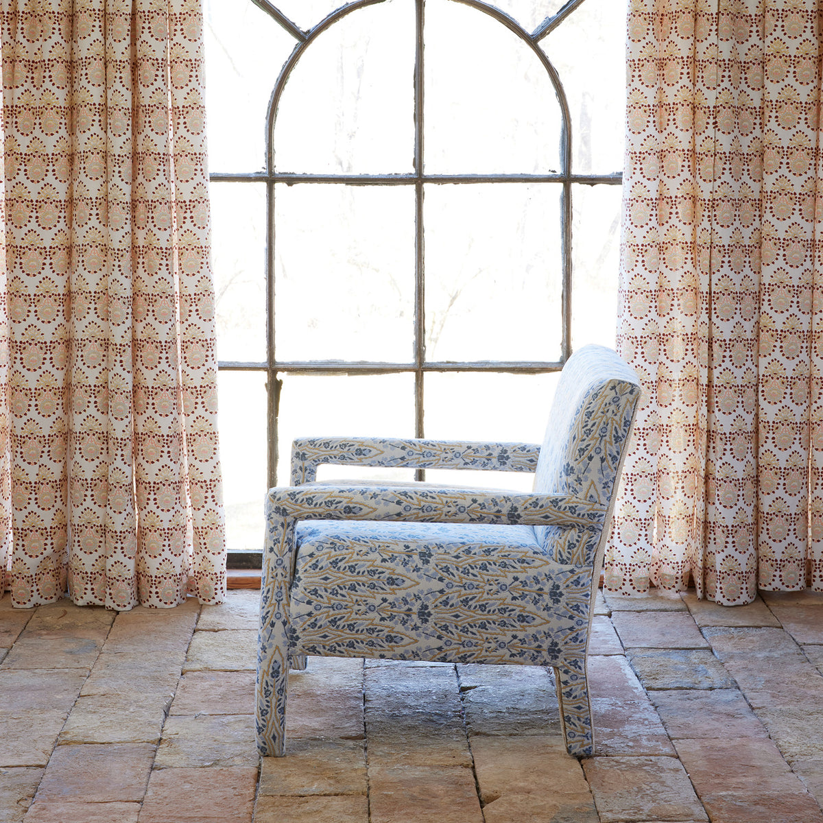 The John Robshaw Square Chair in Marmar Marigold and Natesh Sand, featuring custom upholstery, is positioned in front of a large arched window with patterned curtains and a tiled floor.