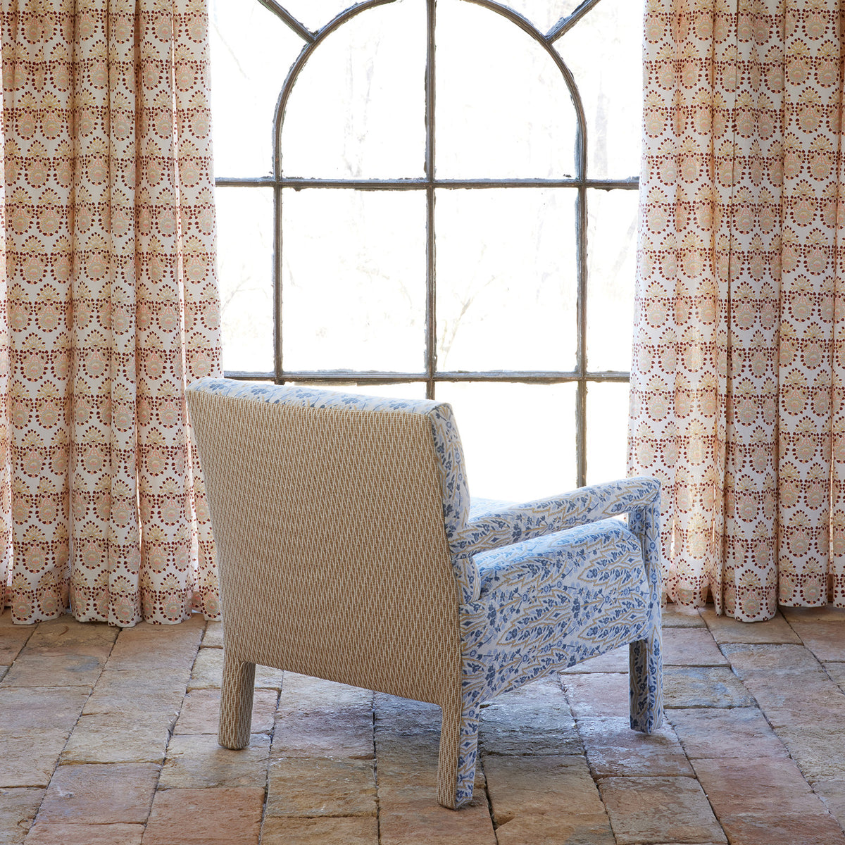 The Square Chair in Marmar Marigold and Natesh Sand by John Robshaw faces a large arched window adorned with patterned curtains, set atop a stone tile floor bathed in natural light. This inviting piece combines stylish design and lasting durability, making it the perfect spot to relax.