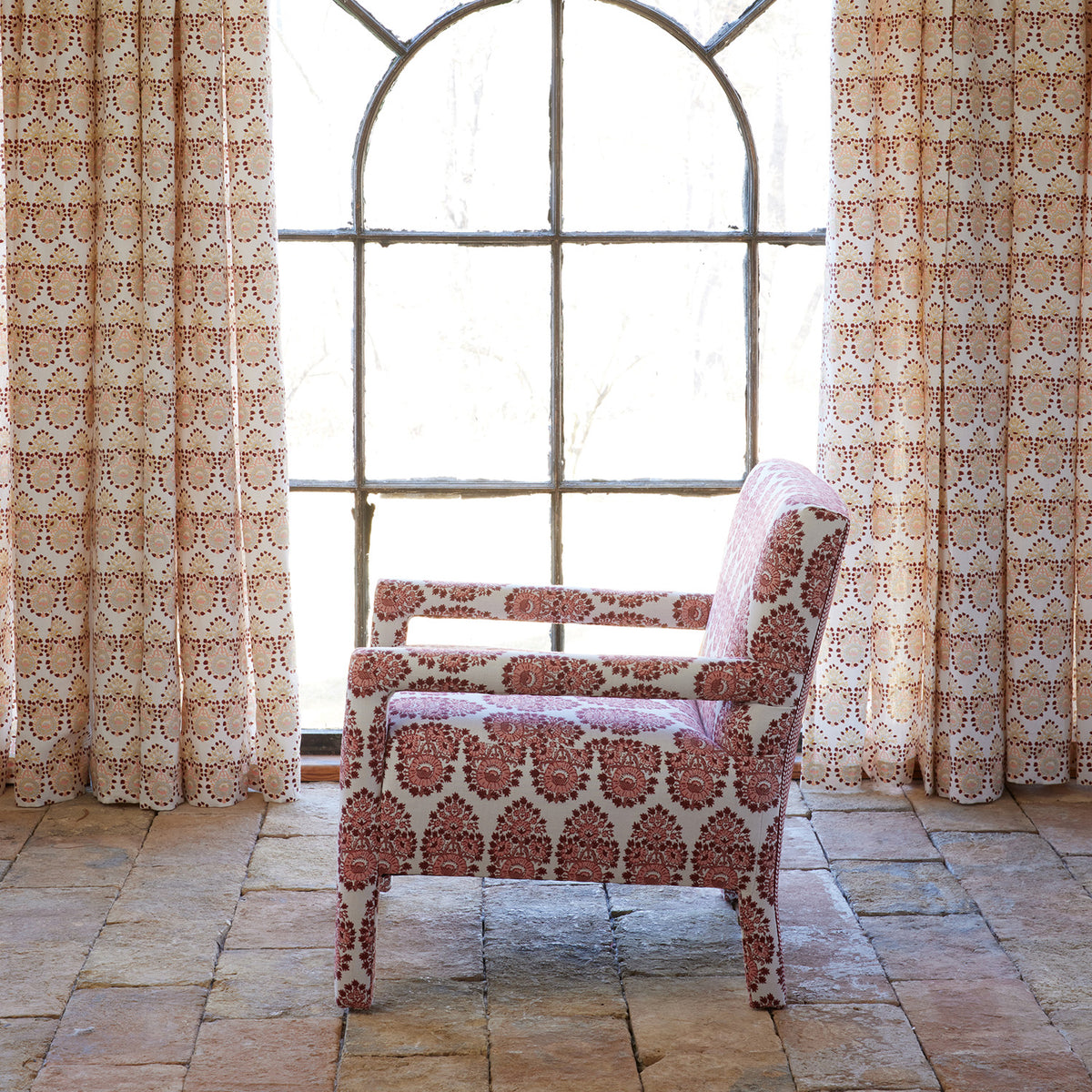 A Square Chair in Lanka Berry and Aleppo Berry by John Robshaw is placed on a tiled floor before a large arched window, surrounded by elegant, patterned curtains made from matching John Robshaw fabrics.