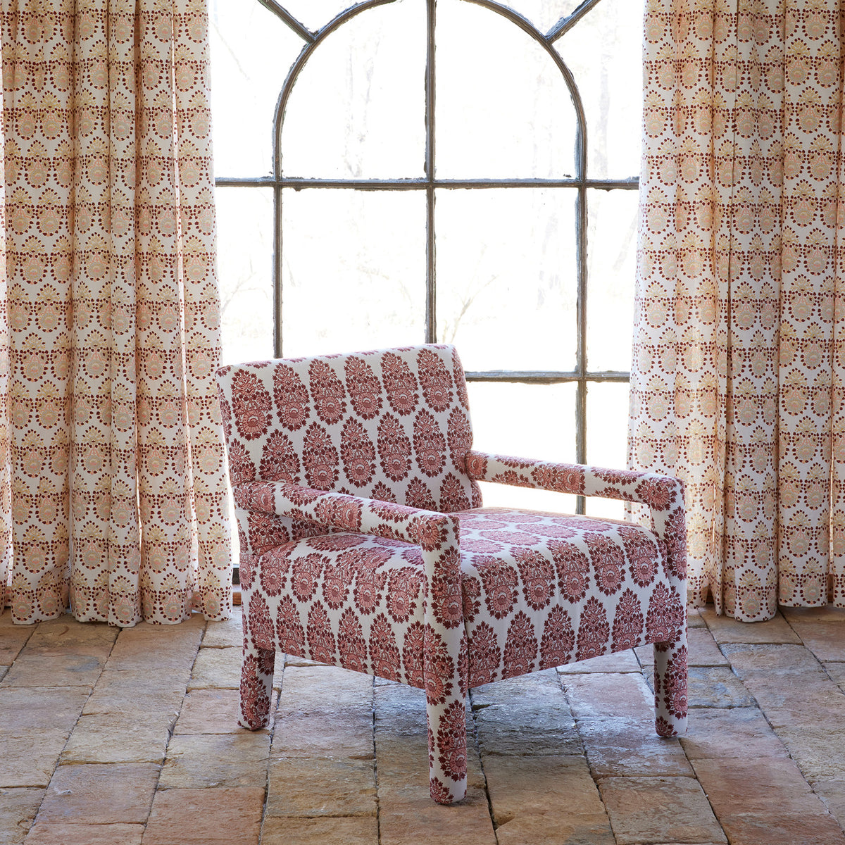 A Square Chair in Lanka Berry and Aleppo Berry by John Robshaw, featuring a red and cream pattern, sits on a tiled floor in front of matching curtains and a large arched window.