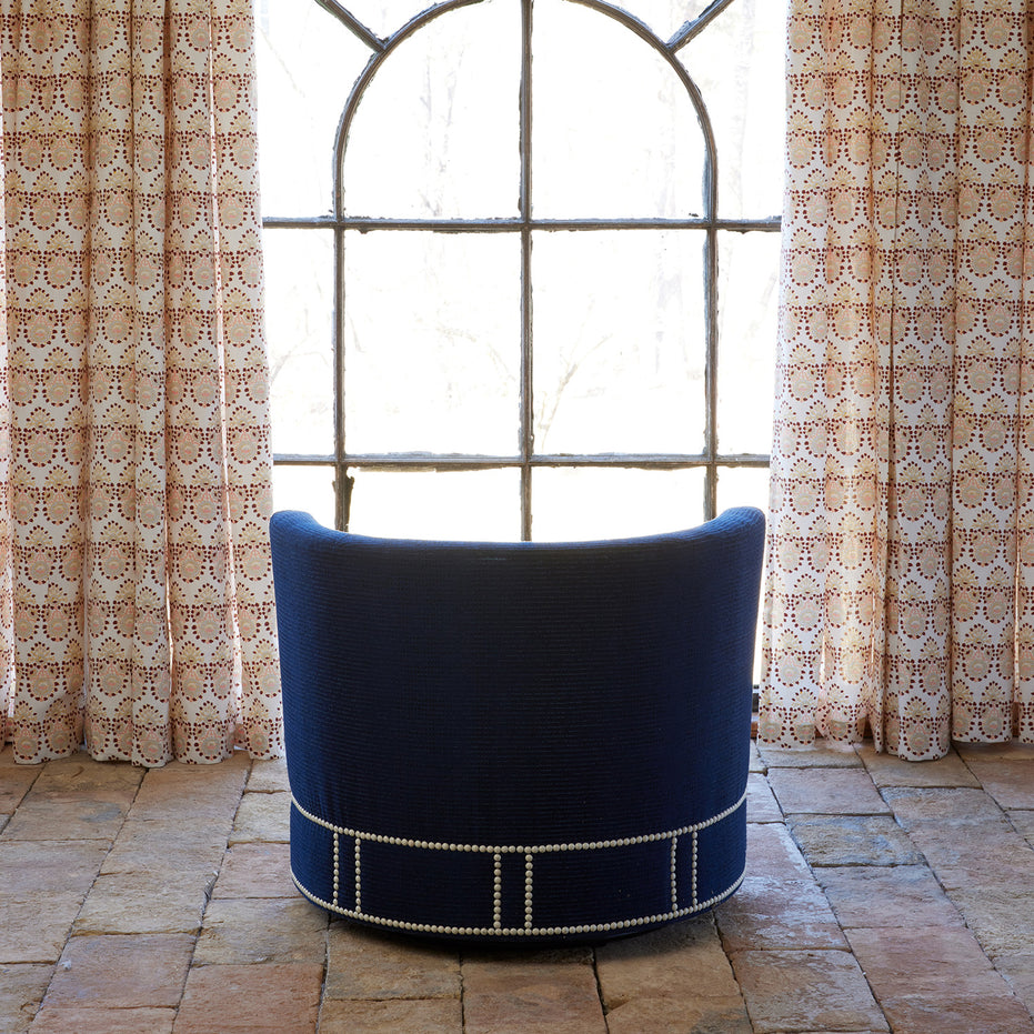 A John Robshaw Round Swivel Chair in Bijal Indigo Performance Fabric, featuring nailhead trim and a swivel base, faces a large arched window, flanked by patterned curtains on a stone tile floor.