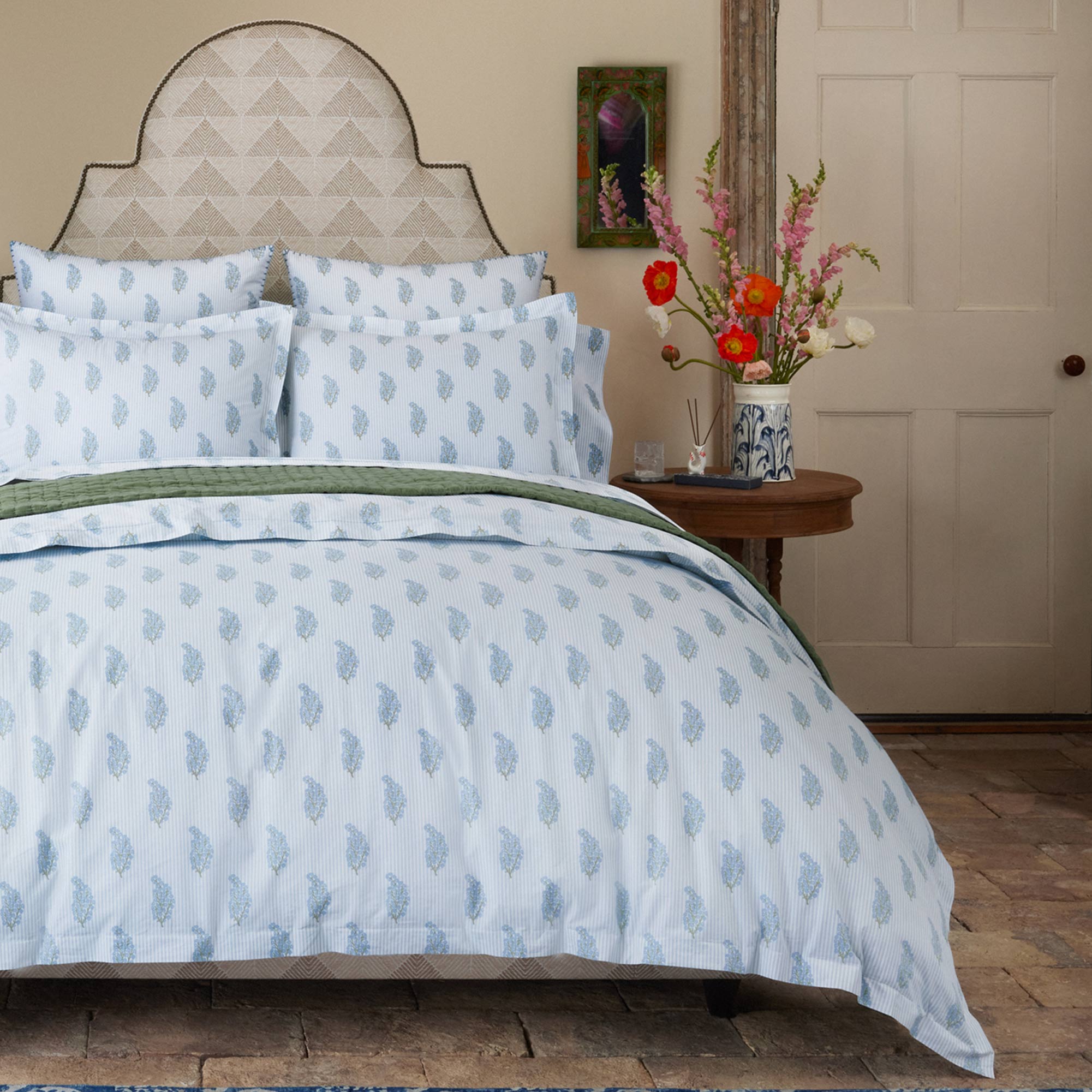 A neatly made bed features the Nirvi Light Indigo Organic Duvet by John Robshaw, showcasing blue and white patterns in organic cotton percale, along with pillows and a headboard. A table with a vase of flowers and a painting on the wall are also visible.