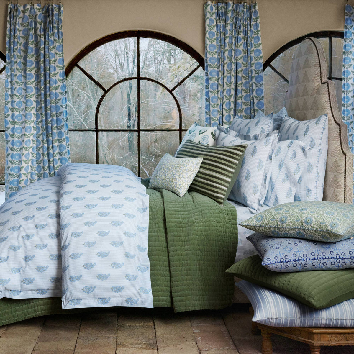 A neatly made bed with John Robshaw Nirvi Light Indigo Organic Sheets, a green quilt, striped pillows, and cushions by large arched windows with blue curtains.