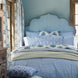 A neatly made bed with a scalloped blue headboard, blue and white floral bedding, and matching curtains in a cozy room with light blue walls and wooden window frames. The Ipsita Indigo Bolster by John Robshaw adds an artisanal touch, perfectly complementing the coastal India vibe of the space.