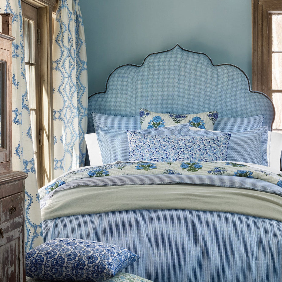 A neatly made bed with a scalloped blue headboard, blue and white floral bedding, and matching curtains in a cozy room with light blue walls and wooden window frames. The Ipsita Indigo Bolster by John Robshaw adds an artisanal touch, perfectly complementing the coastal India vibe of the space.