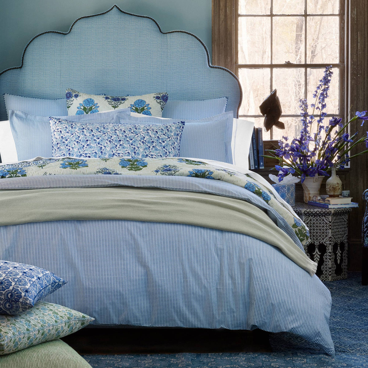A neatly made bed features the John Robshaw Cinde Navy Organic Duvet with blue and white bedding, paired with floral pillows and a patterned headboard; a vase of purple flowers rests on a side table by the window.