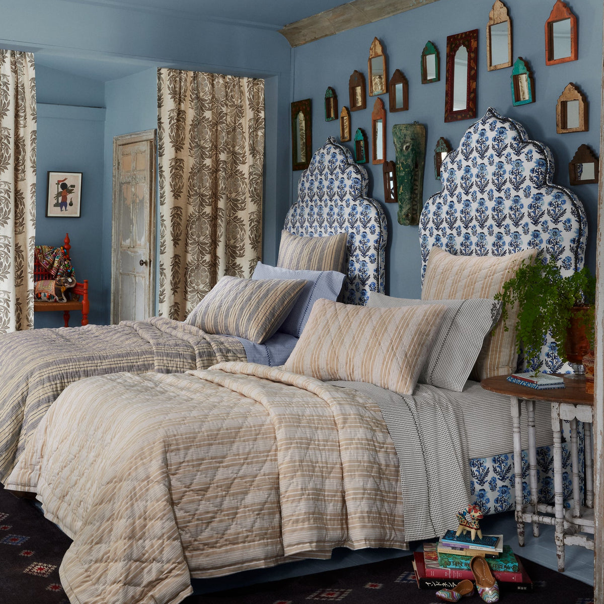 Two twin beds with patterned headboards and striped bedding in a blue room, featuring wall art, a small side table, and curtains crafted from John Robshaw’s Hina Azure Fabric—a delicate floral design inspired by traditional block printing.
