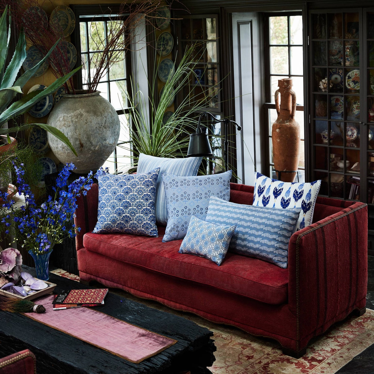 A red velvet sofa adorned with an assortment of blue and white patterned pillows—including the John Robshaw Aanan Light Indigo Pillow—alongside a hand block printed blue floral cushion and an embroidered pillow, sits in a richly decorated room filled with plants, pottery, and dark wooden furniture near large windows.