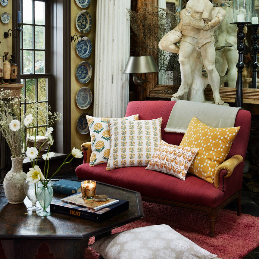 A cozy living room featuring a vibrant red sofa adorned with the John Robshaw Anisha Marigold Pillow, its hand block printed yellow and white floral patterns adding rustic charm, alongside a large statue, decorative plates on the wall, and a table styled with flowers—perhaps marigolds—a candle, and books. The inviting space is beautifully accented by these artisanal textiles.