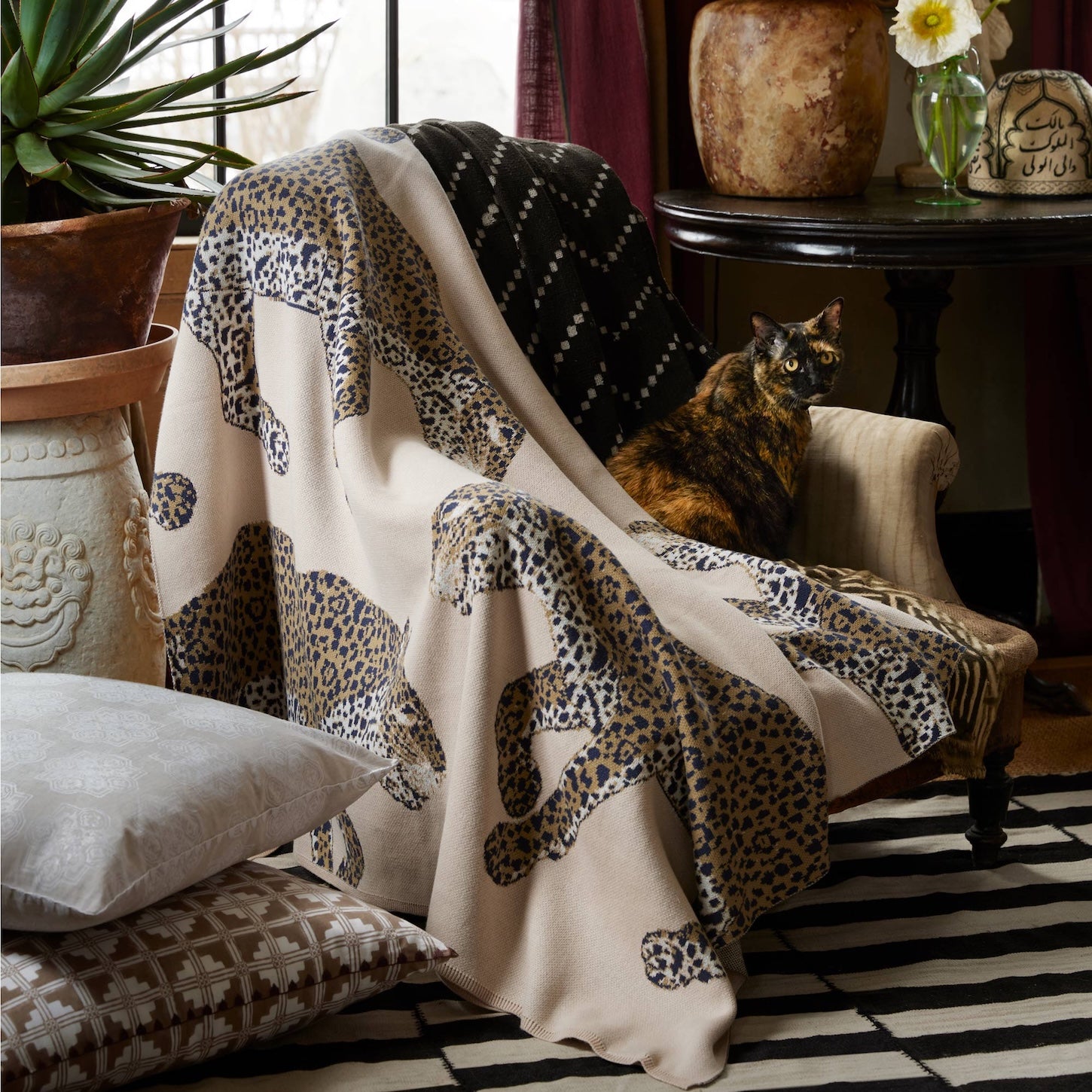 A tortoiseshell cat sits on a beige armchair draped with a leopard-print blanket, surrounded by patterned pillows—including the John Robshaw Yasha Decorative Pillow—a rug, and a round table with flowers and pottery.