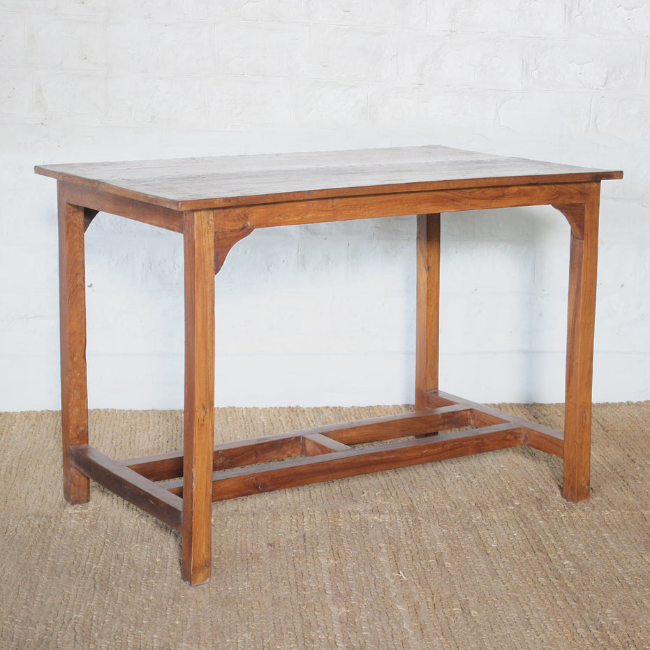 A rectangular, hand crafted Taru Teak Table by John Robshaw with a flat top and simple frame, placed on a woven rug against a white brick wall.