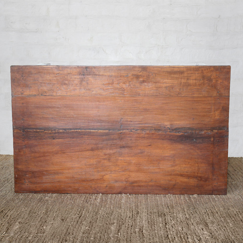 Introducing the Taru Teak Table by John Robshaw—a beautifully handcrafted rectangular wooden tabletop featuring visible grain and a central seam. Set on a textured brown floor against a white brick wall, this piece is perfect for vintage furniture enthusiasts.