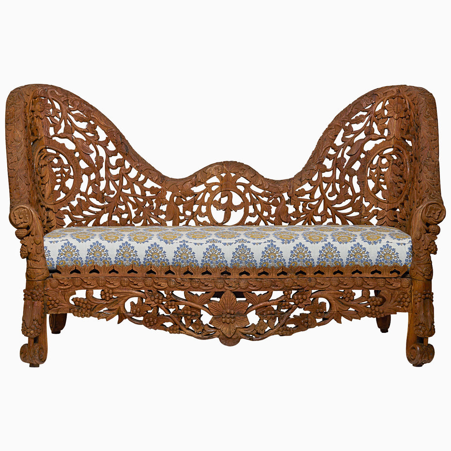 The Candra Teak Sofa by John Robshaw features intricate floral carvings on rich teak and a cushioned seat upholstered in luxurious blue and gold patterned fabric, beautifully capturing the timeless elegance of an Anglo Indian sofa.