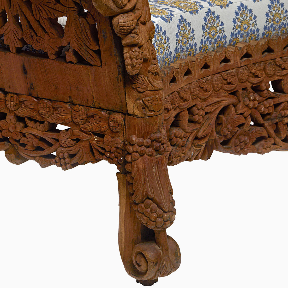 Close-up of an intricately carved leg from the Candra Teak Sofa by John Robshaw, reminiscent of vintage furniture with floral and grapevine motifs, paired with a blue and gold patterned upholstered seat.