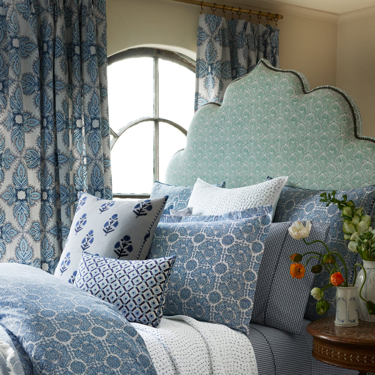 A bed featuring a patterned light blue headboard, blue and white pillows and bedding crafted from elegant Divit Willow Fabric by John Robshaw, complemented by floral curtains and a round window behind, with a vase of flowers resting on a nearby table.