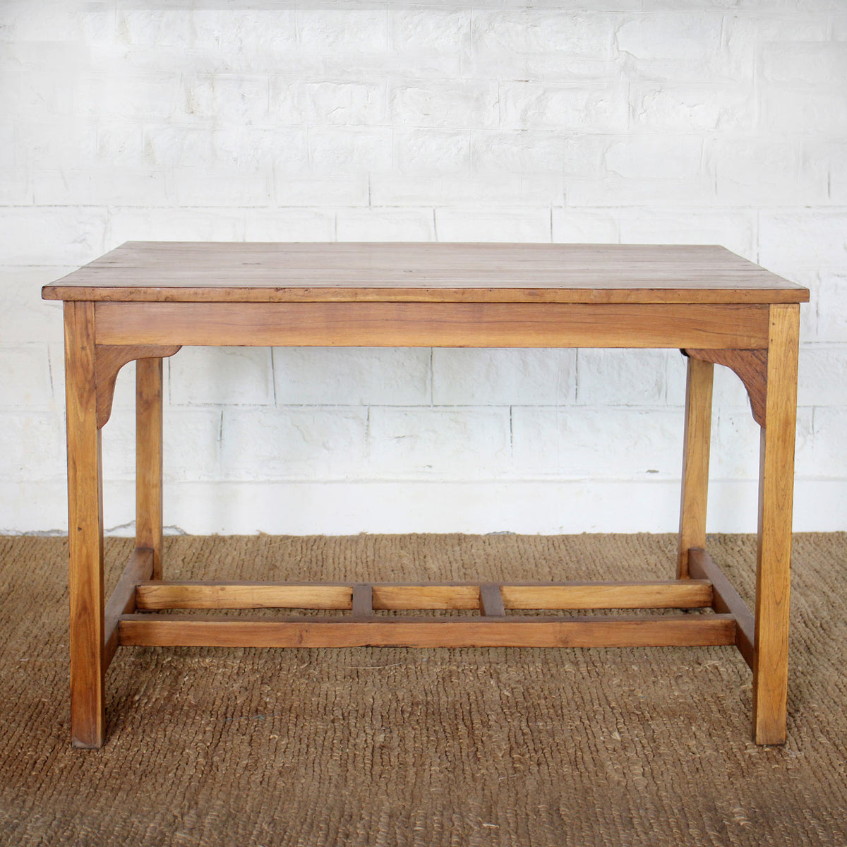 The Taru Teak Table by John Robshaw, hand crafted with a rectangular solid teak top and simple, straight legs, is displayed on a woven rug against a white brick wall.