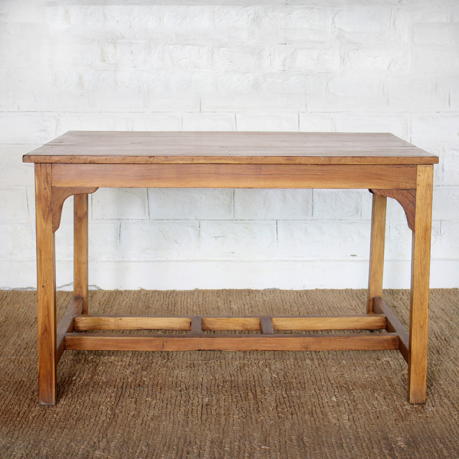 The Taru Teak Table by John Robshaw, hand crafted with a rectangular solid teak top and simple, straight legs, is displayed on a woven rug against a white brick wall.