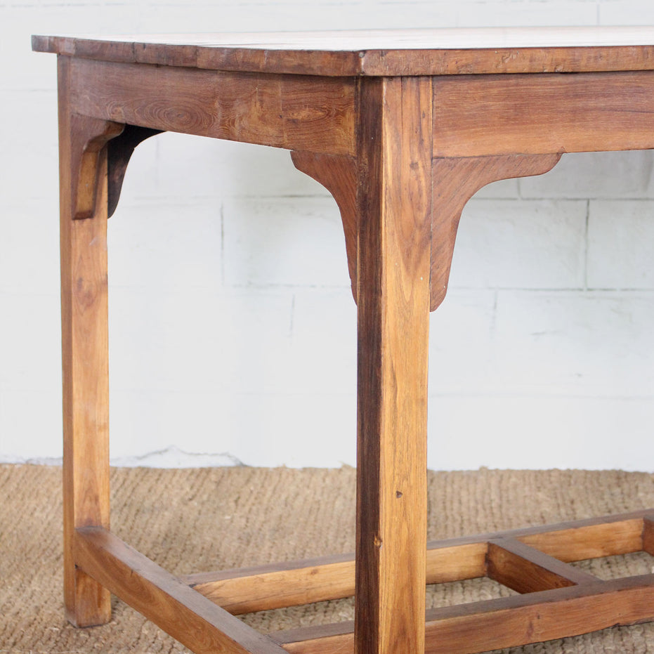 A close-up of the John Robshaw Taru Teak Table, featuring hand-crafted solid teak construction with rectangular legs and corner brackets, placed on a woven rug against a white brick wall.