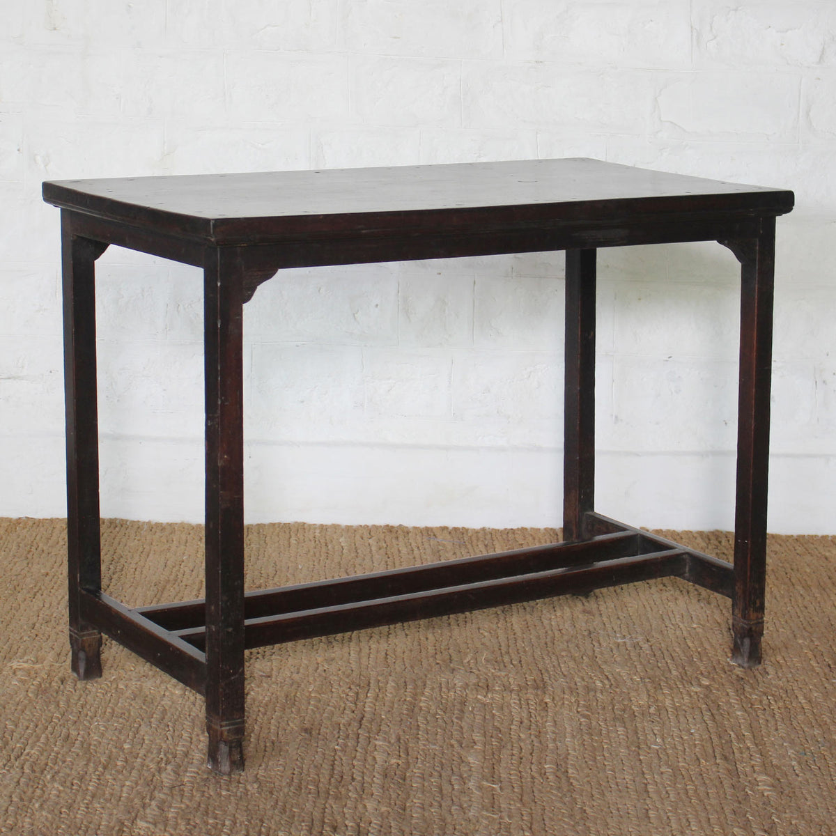 The Chhaya Teak Table by John Robshaw, a handcrafted rectangular dark wood piece featuring four straight legs and a sturdy stretcher base, sits atop a woven mat against a white brick wall, radiating the timeless charm of vintage furniture.