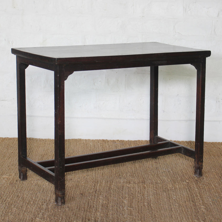 The Chhaya Teak Table by John Robshaw, a handcrafted rectangular dark wood piece featuring four straight legs and a sturdy stretcher base, sits atop a woven mat against a white brick wall, radiating the timeless charm of vintage furniture.